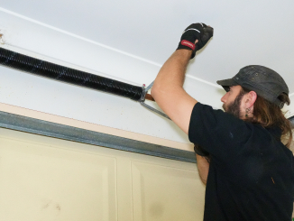 A man in a black shirt and cap is adjusting a garage door spring, using tools to tighten the large black spring above the door. He wears protective gloves and focuses on the task.